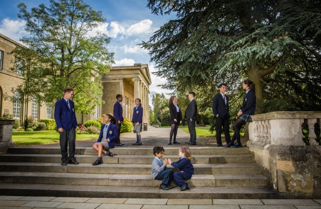 Students at Mill Hill School gather outside on the steps, wearing uniforms and engaging in conversation, set against the backdrop of a historic school building and greenery on a sunny day.