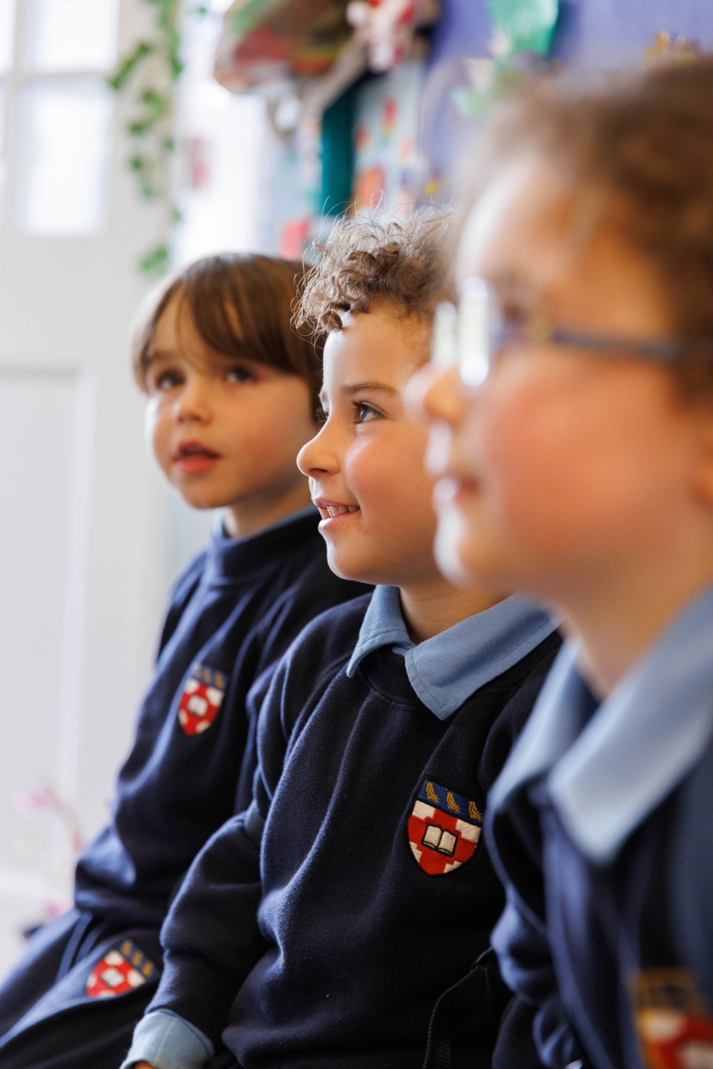 Three young children in school uniforms sit together, focused and engaged, during a classroom activity, with a colourful and creative environment in the background.