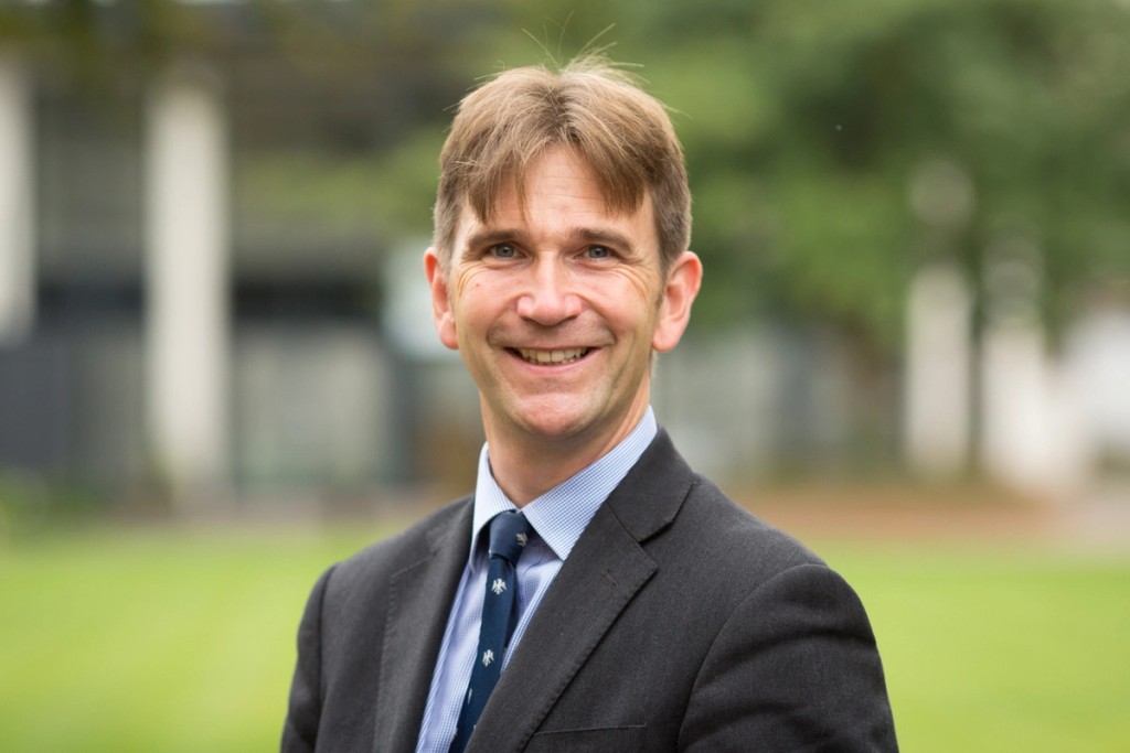 Portrait of the Chief Executive Officer of the Mill Hill Education Group, smiling in a formal suit and tie, standing outdoors with a green, blurred background.