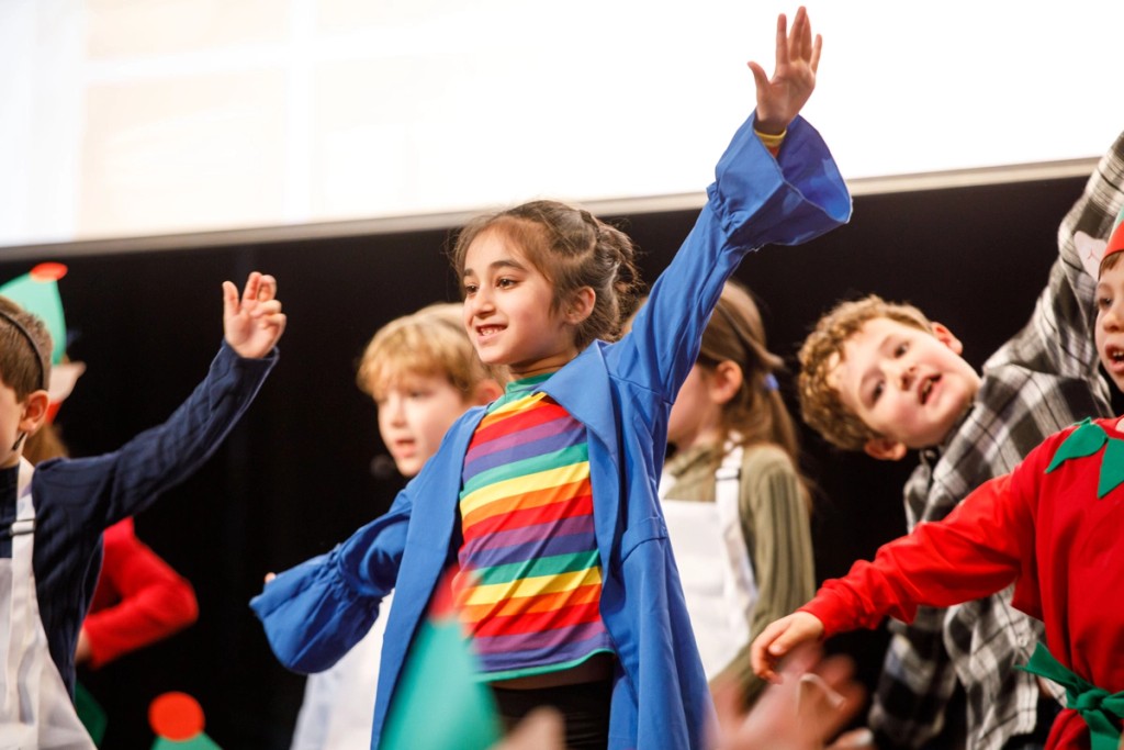 Children on stage at Mill Hill Pre-Prep, performing in colourful costumes with joyful expressions, raising their hands during a school play or performance.