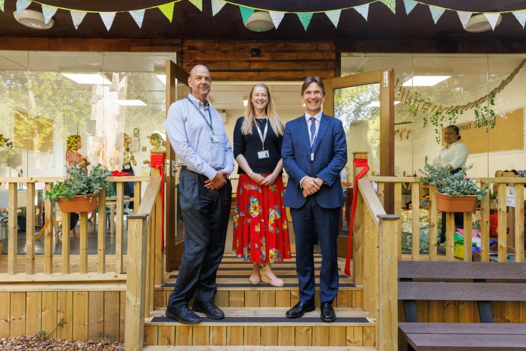 Three staff members at Mill Hill Pre-Prep stand on the wooden steps of the school entrance, smiling and posing together in front of a brightly decorated classroom, with bunting hanging above them.