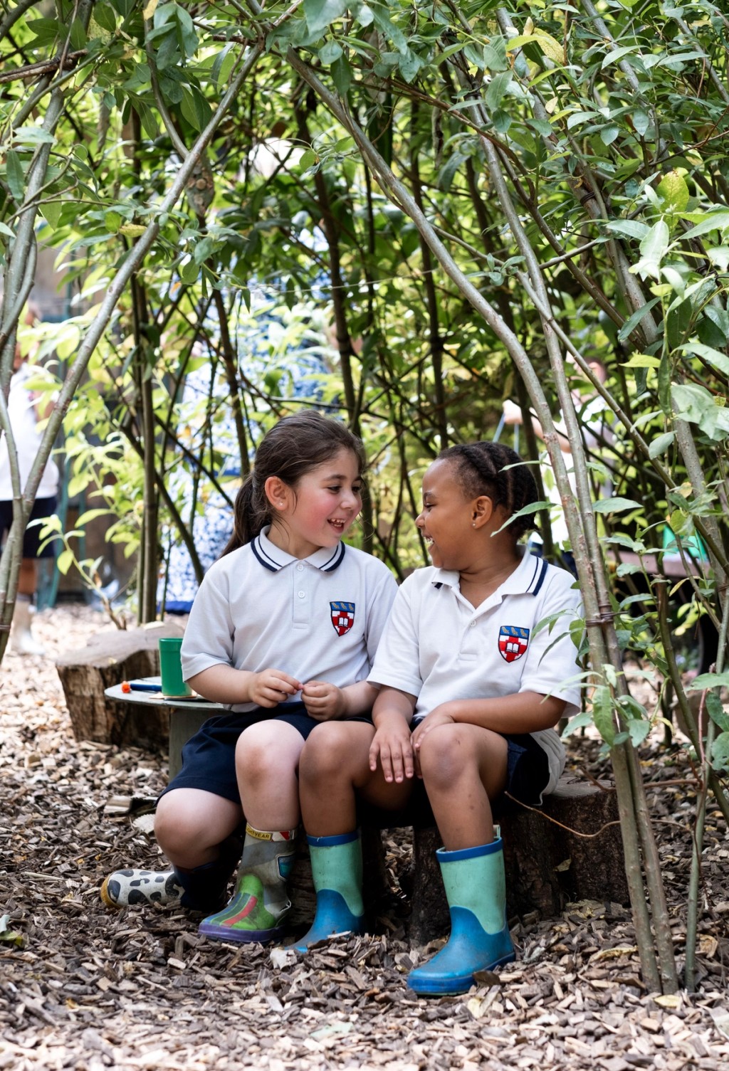 Two children in Mill Hill Pre-Prep uniforms sit together in an outdoor learning area surrounded by greenery, smiling and chatting while wearing wellies, enjoying nature-based activities.