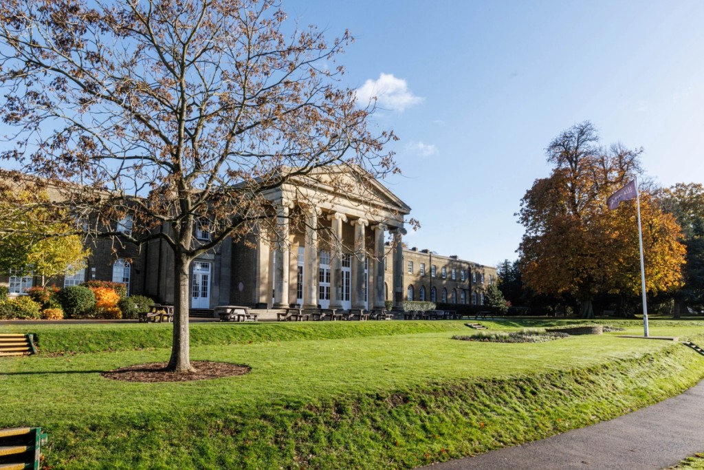 Exterior view of Mill Hill School's main historic building, set on a spacious green lawn with autumn trees and a clear blue sky.