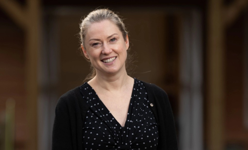 Portrait of the Head of Grimsdell at Mill Hill School, smiling and standing outdoors, wearing a black polka dot dress and cardigan with a warm wooden background.