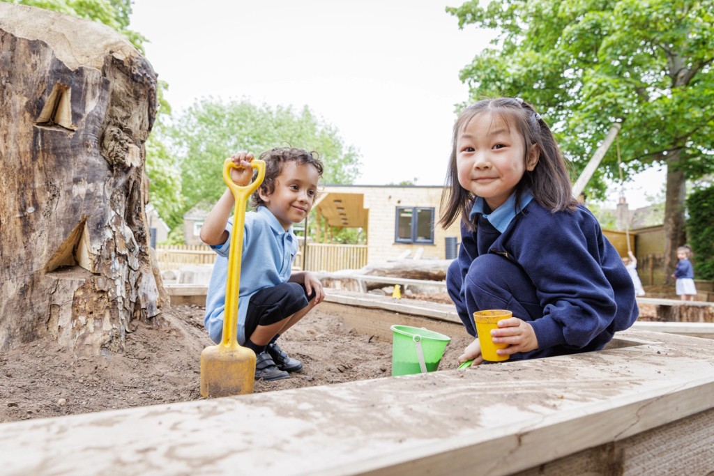 Two children play in a sandbox with a shovel and buckets. Trees and a building are in the background.