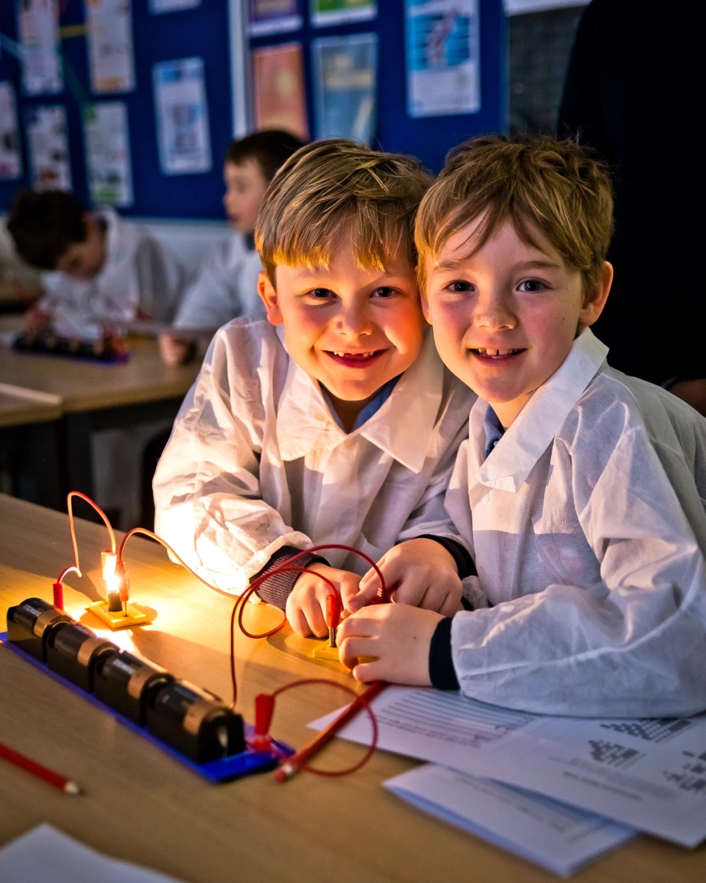 Two children in a classroom at Mill Hill Pre-Prep, wearing lab coats and working on a simple electrical circuit with light bulbs, smiling and engaged in a science activity.