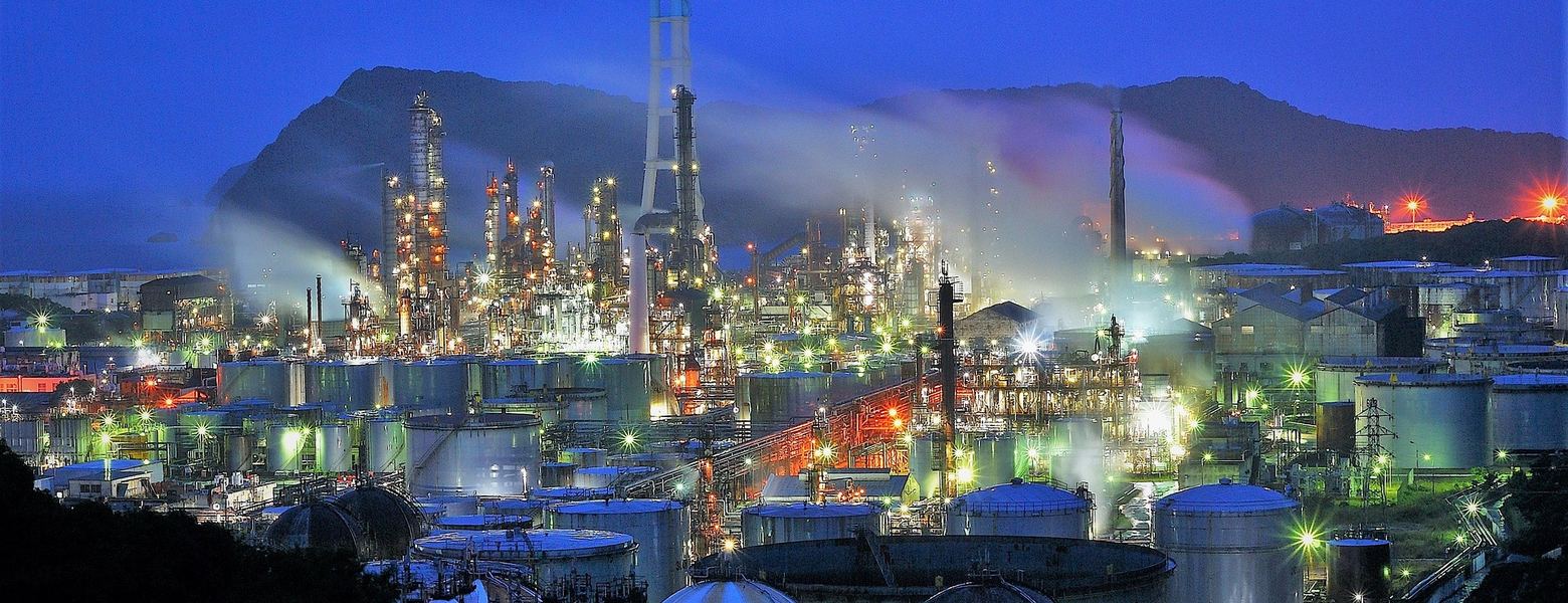 A refinery complex lit up at dusk below a mountain ridge