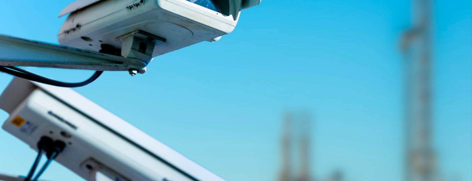Two security cameras mounted on a pole, with industrial storage tanks blurred behind