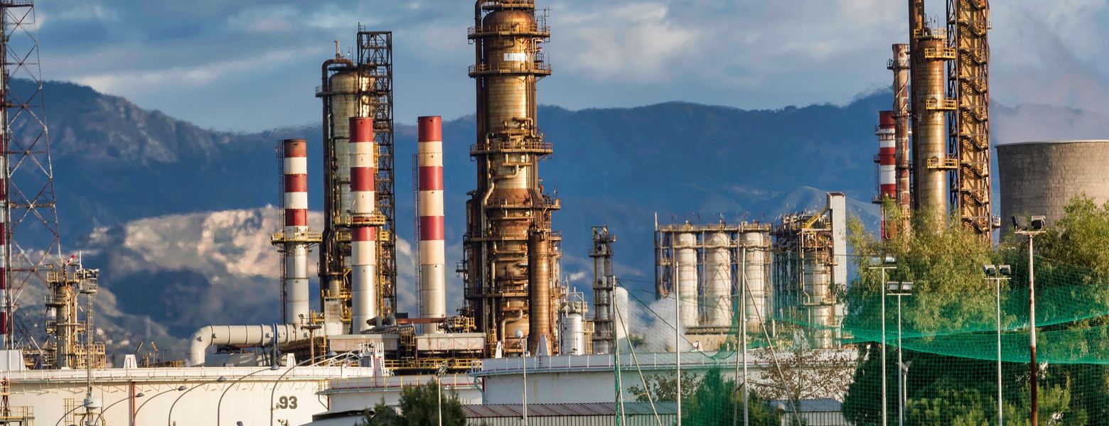 A refinery with distillation columns and striped stacks below a mountain ridge