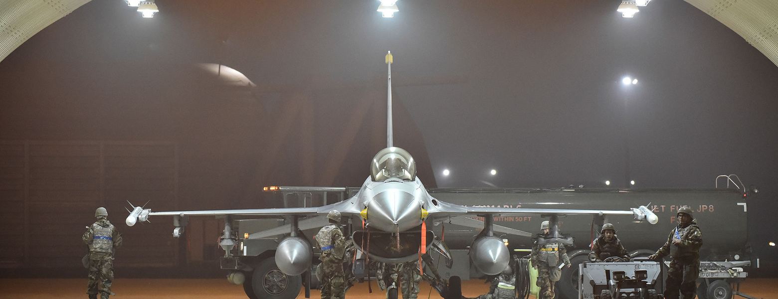 Ground crew standing beside a fighter jet inside a lit hangar at night