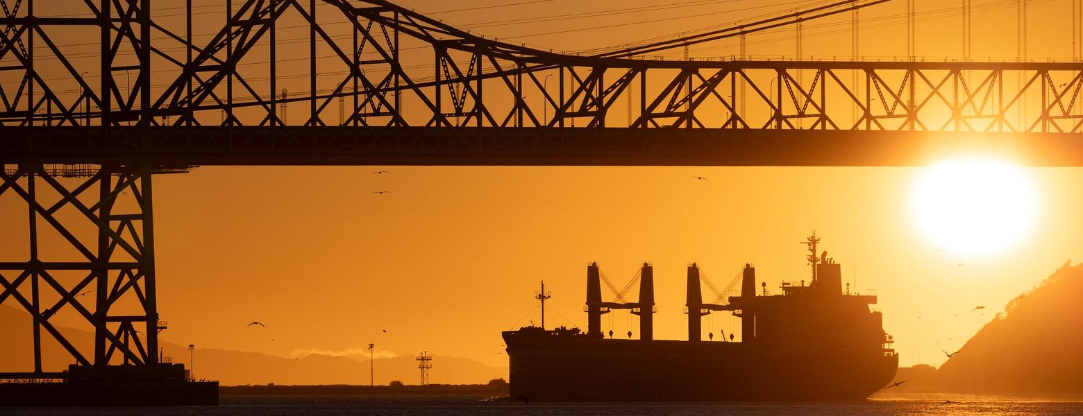 A cargo ship silhouetted beneath a steel bridge at sunset