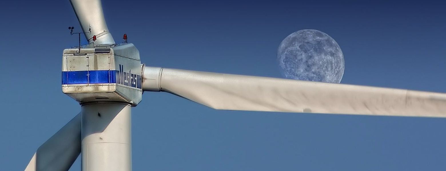 The nacelle and blade of a wind turbine against a blue sky, with the moon behind