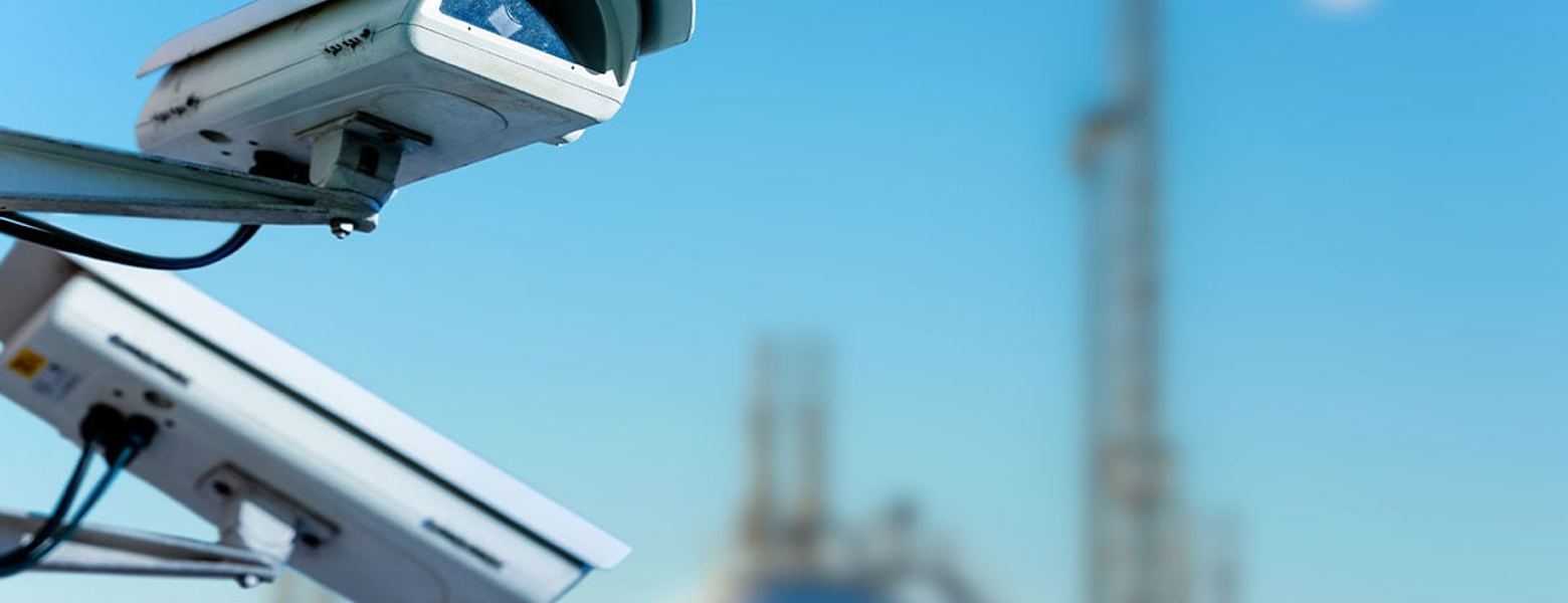 A security camera mounted above an industrial site, with refinery stacks blurred behind it