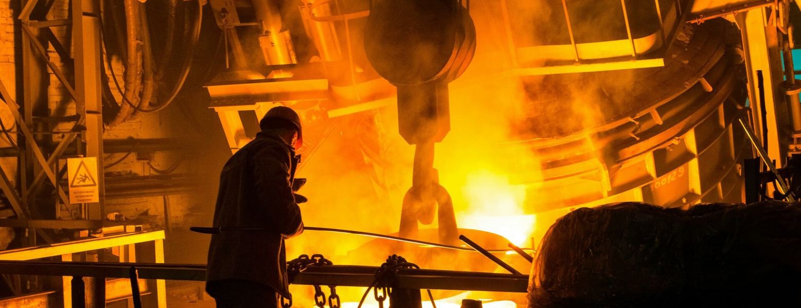 A worker beside a crane hook and ladle in the glow of a steel foundry
