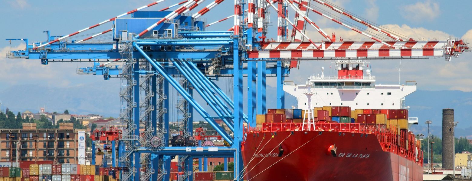 Gantry cranes and stacked containers at a port, with a container ship berthed alongside