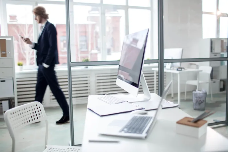 Moderne kontor med dataskjermer og dokumenter på pulten I bakgrunnen går en eiendomsmegler i dress mens han ser på mobilen sin