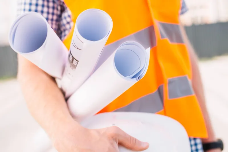 En entreprenør iført oransje refleksvest holder sammenrullede byggetegninger og en hjelm klare for et byggeprosjekt