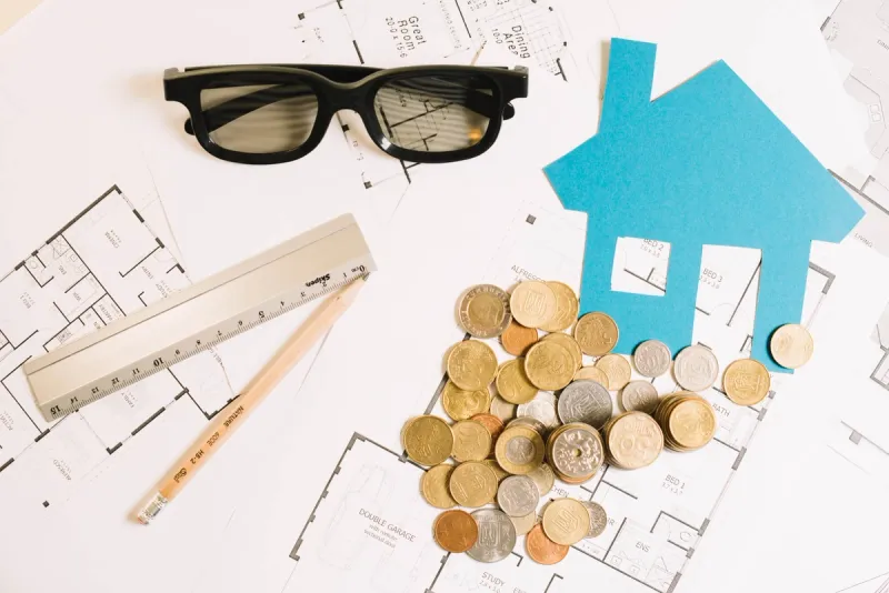 Arkitekttegninger med mynter linjal blyant og et blått papirutklipp av et hus symboliserer kostnader for takarbeid