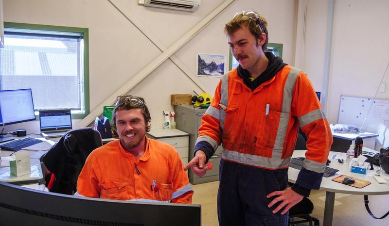 Two engineering workers in orange high-visibility clothing reviewing information on a computer in a workplace office.