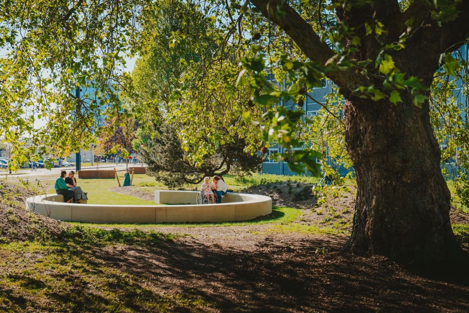 Patiënten en bezoekers ontspannen op een ronde zitbank in de tuin van UZ Gent, onder meer iemand in een rolstoel.