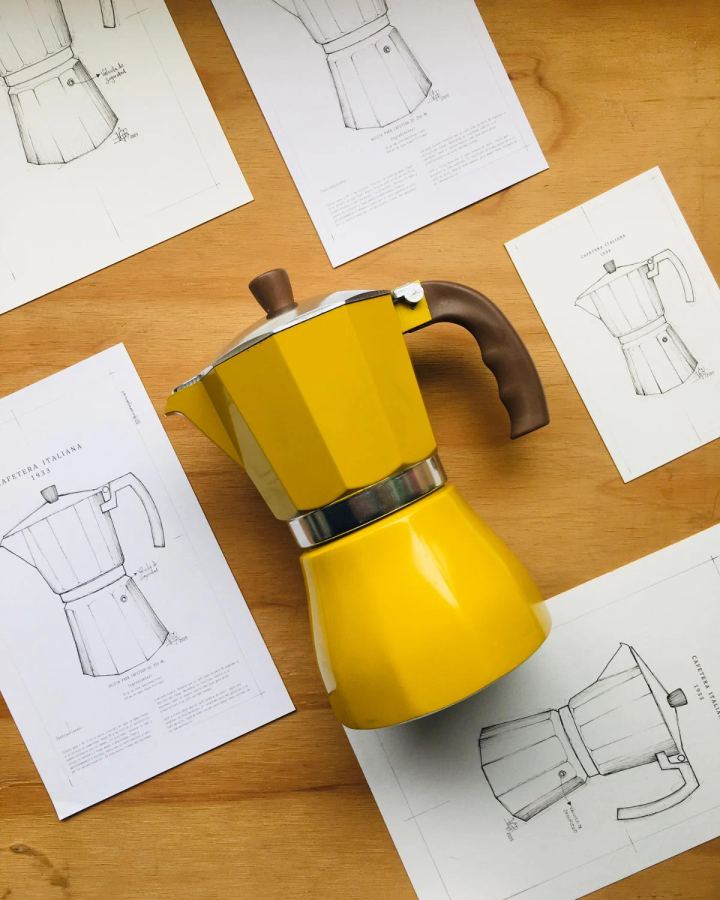 Cafetera italiana amarilla rodeada de bocetos a lápiz sobre una mesa de madera
