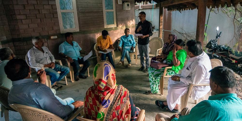 A community meeting in a rural setting with several people seated in plastic chairs arranged in a circle.