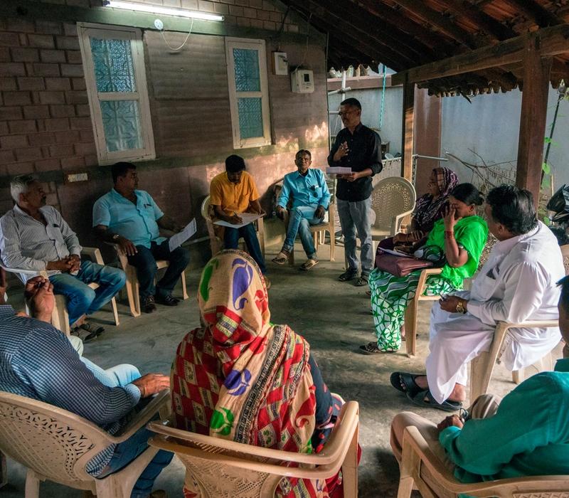 A group of people seated in a semi-circle under a shaded veranda during a community meeting; one person stands and speaks.