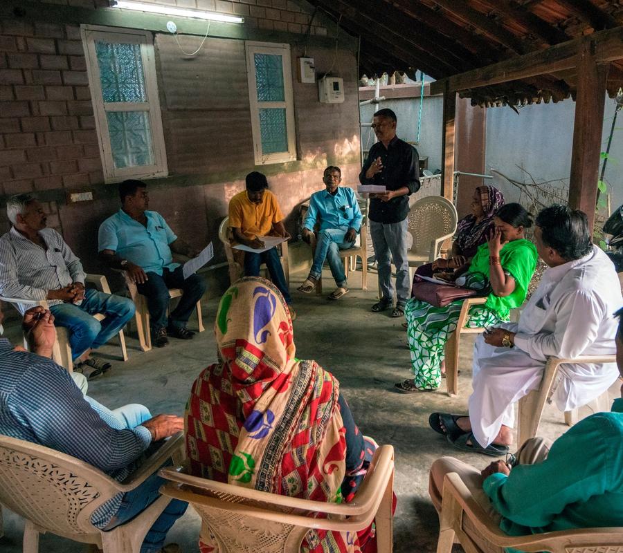 A community meeting in a rural setting with several people seated in plastic chairs arranged in a circle.