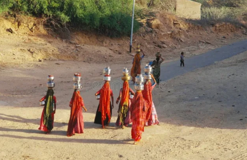 An elevated rear view of a group of women walking while carrying multiple water vessels on their heads and hips.