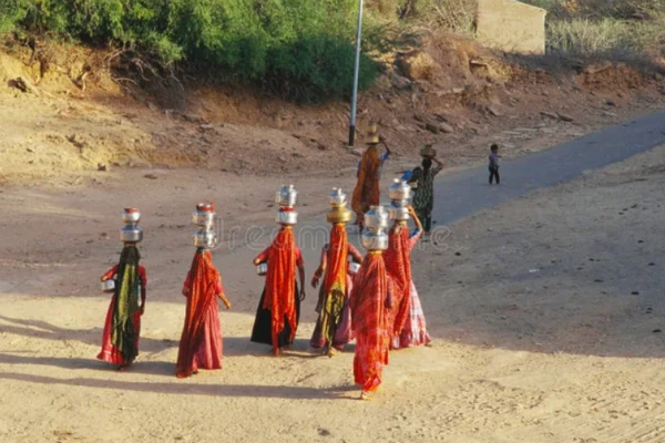 An elevated rear view of a group of women walking while carrying multiple water vessels on their heads and hips.