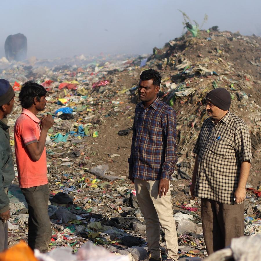 Four men stand in a landfill filled with scattered trash. Two cows graze in the background under a hazy sky.