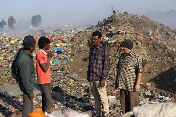 Four men stand and talk in a large landfill filled with piles of garbage and plastic waste.