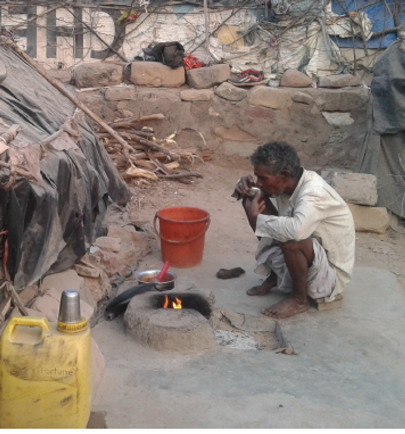 A migrant worker sits crouched outside his hut, drinking tea beside a burning chulha. 