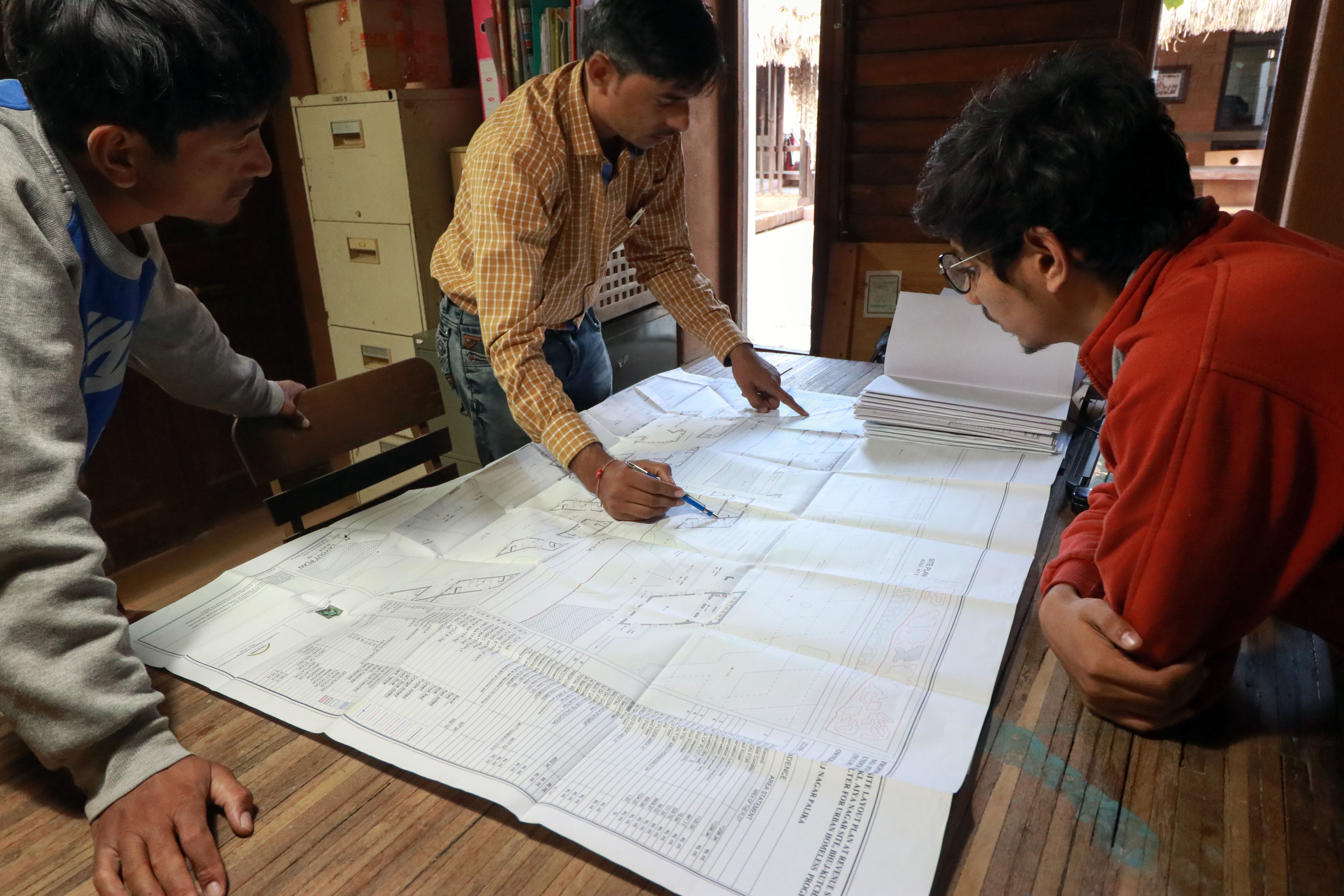 Three men examine a large blueprint on a table; one points at a detail during a discussion in an office.