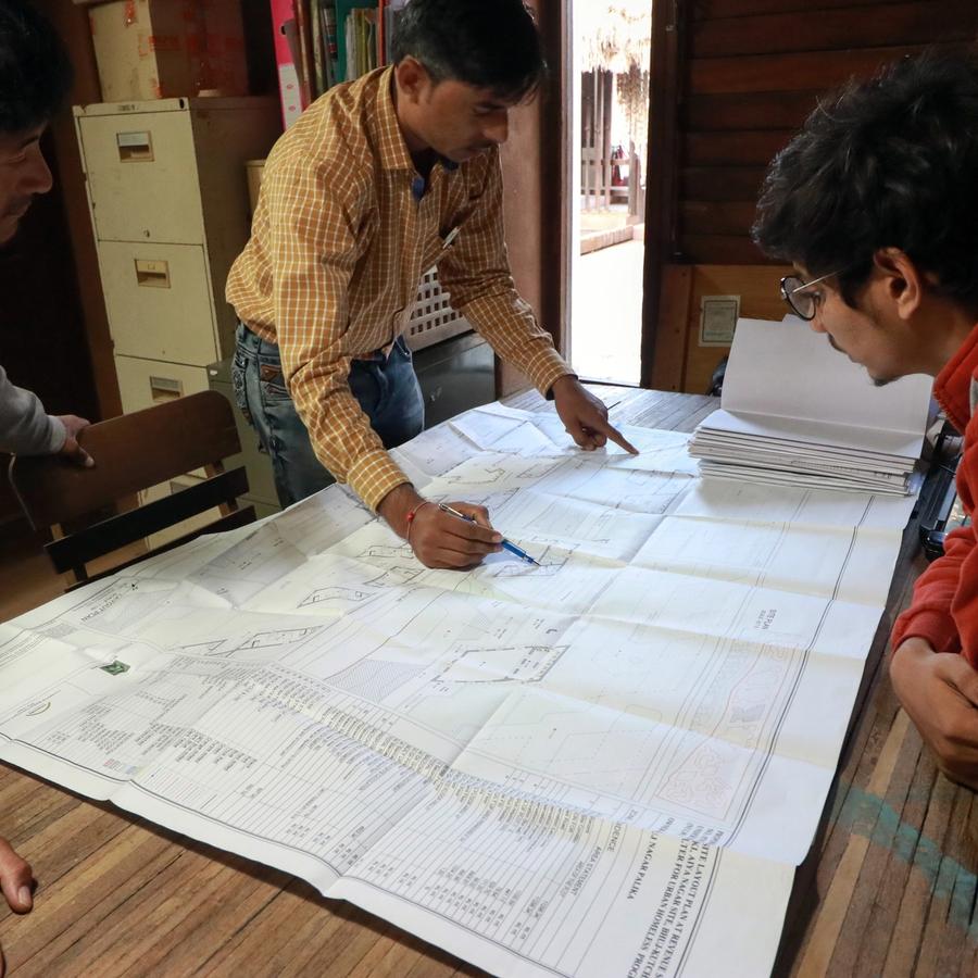 Three men examine a large blueprint on a table; one points at a detail during a discussion in an office.