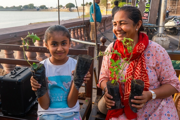 A mother and her daughter sit on a bench, smiling and holding saplings in their hands