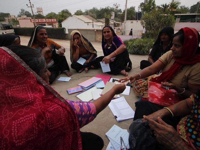 A woman sits on a chair, surrounded by people, writing in a notebook on her lap.