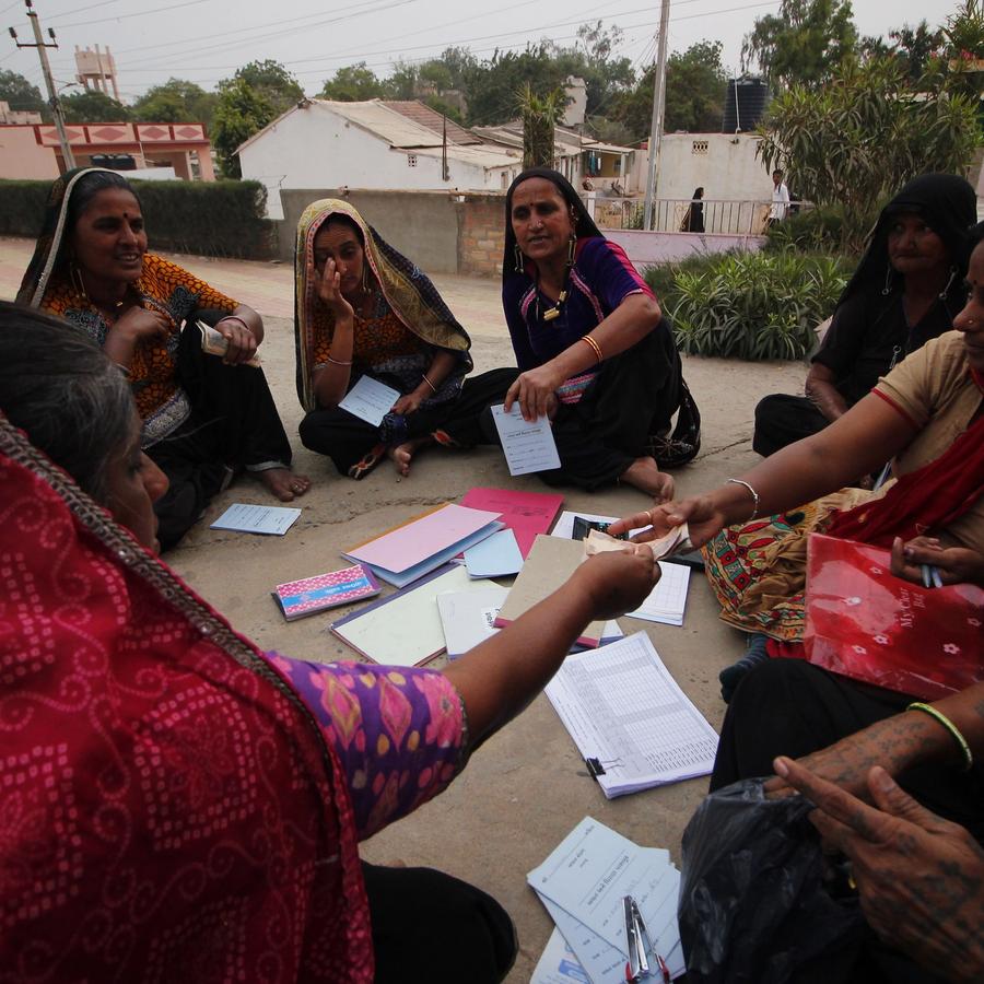 A group of women in colorful traditional attire sit in a circle on the ground, exchanging documents and money.