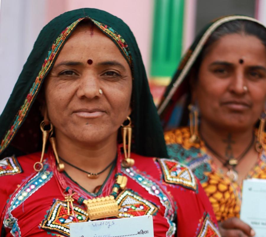 A woman in traditional colorful clothing and jewelry holds up a document written in Gujarati.