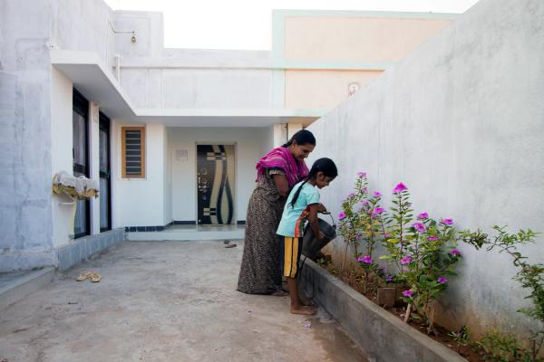 A young girl and an older woman watering the plants in their house. A white single storey house in the background.