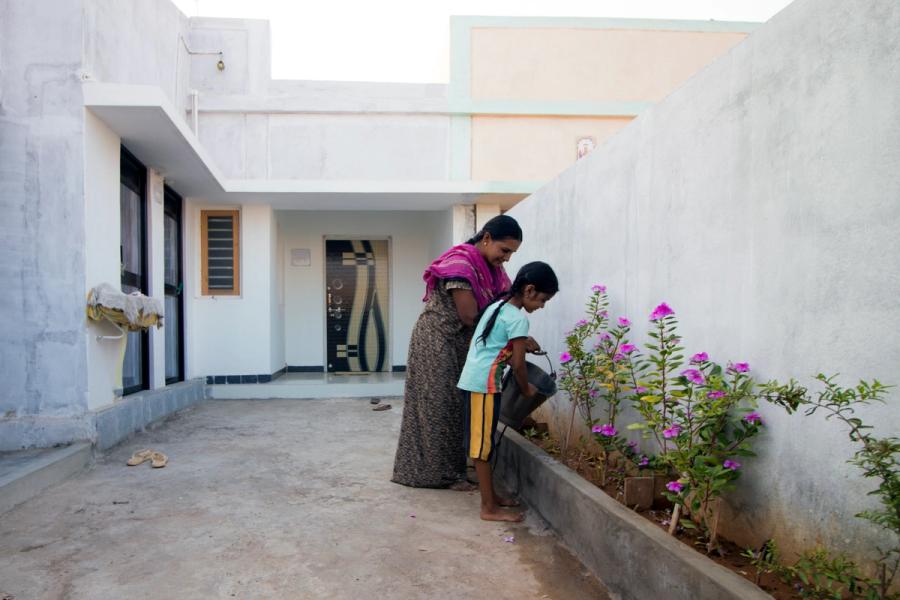 A young girl and an older woman watering the plants in their house. A white single storey house in the background.