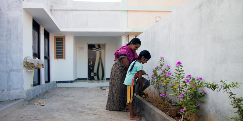 A young girl and an older woman watering the plants in their house. A white single storey house in the background.