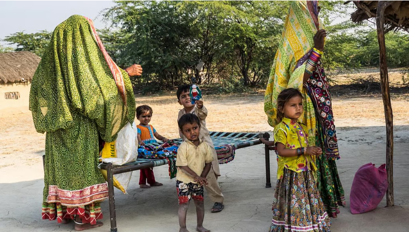 Courtyard of a hut. 4 Kids and 2 women are standing near a bed. Kids are playing. 