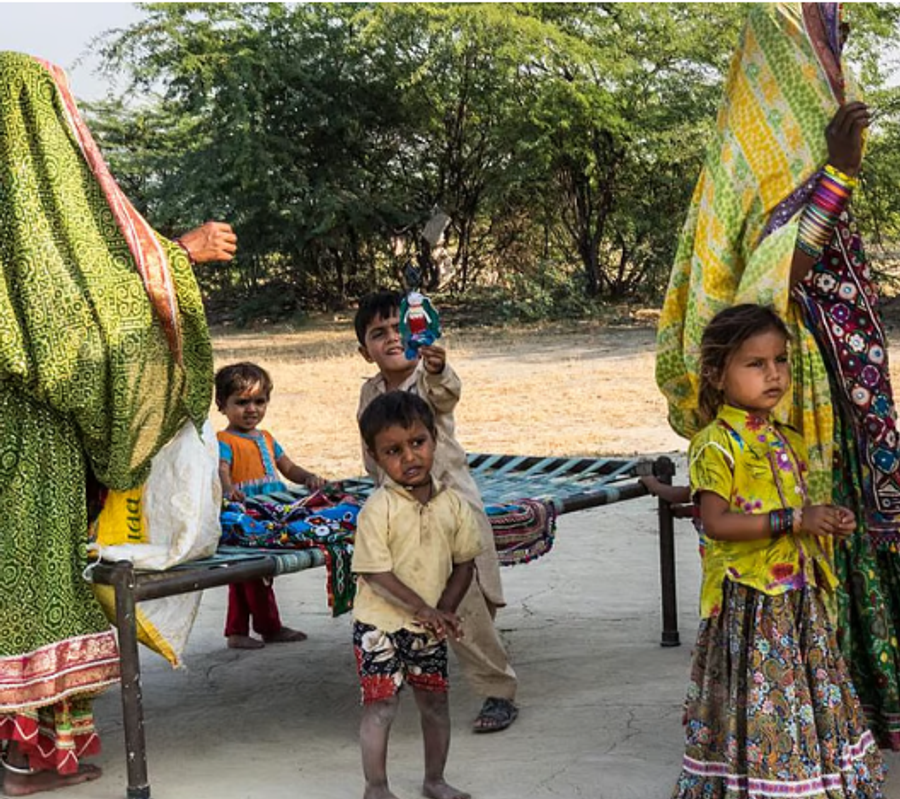 Courtyard of a hut. 4 Kids and 2 women are standing near a bed. Kids are playing.