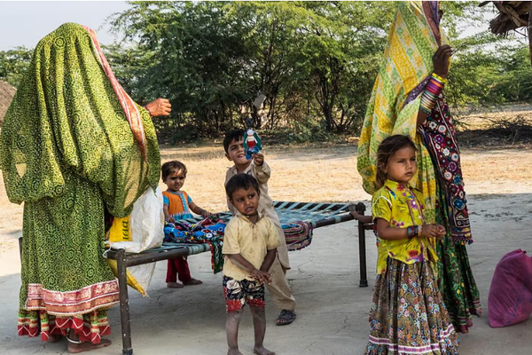 Courtyard of a hut. 4 Kids and 2 women are standing near a bed. Kids are playing.