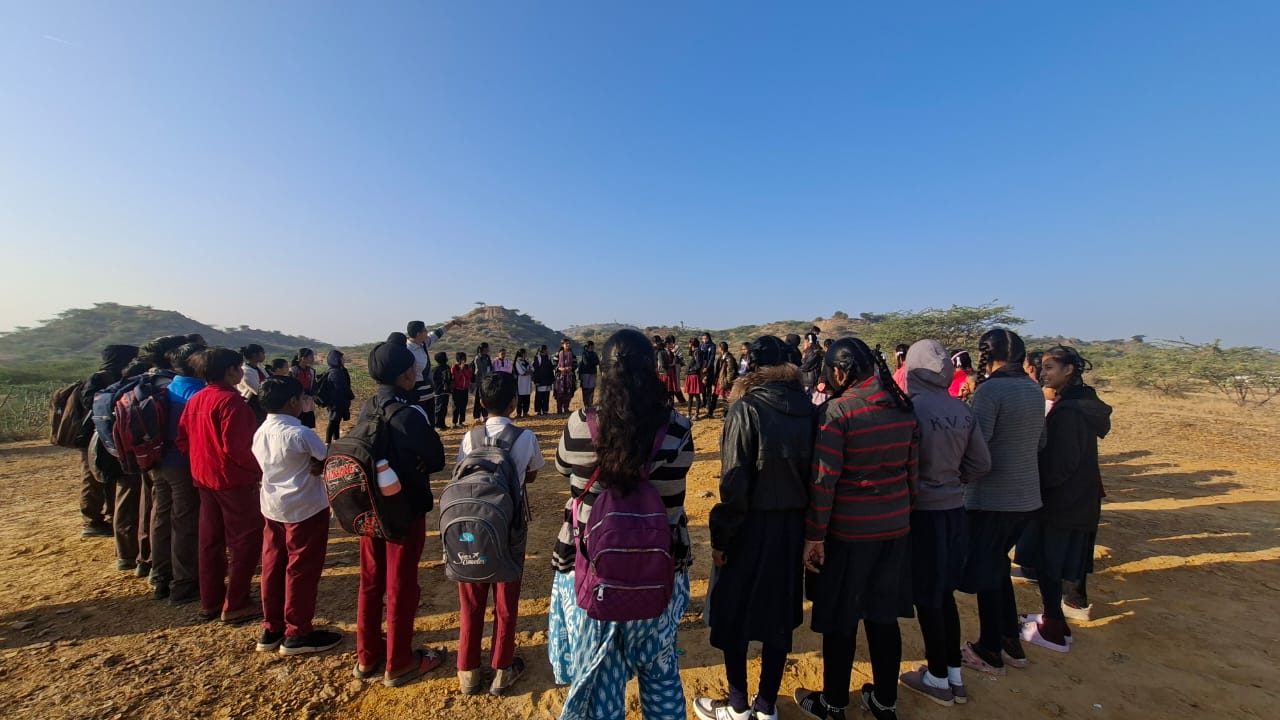 Children standing in circle in an open ground. A man is pointing to a far away hill standing in centre