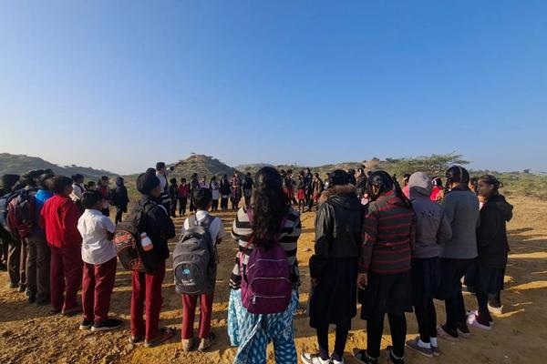 Children standing in circle in an open ground. A man is pointing to a far away hill standing in centre