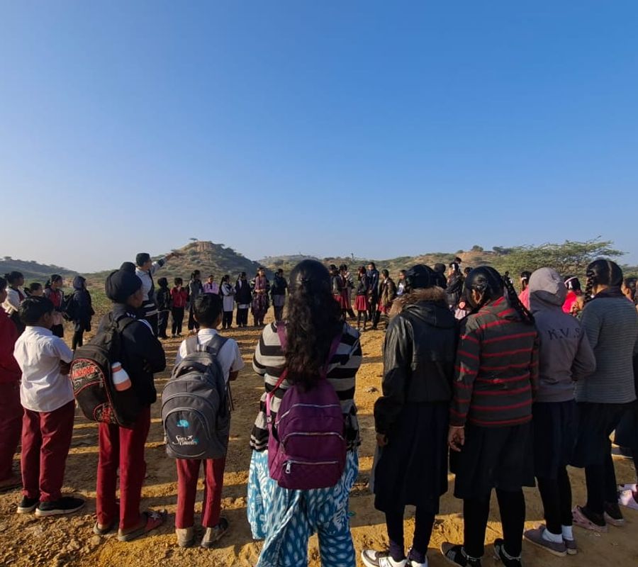 Children standing in circle in an open ground. A man is pointing to a far away hill standing in centre
