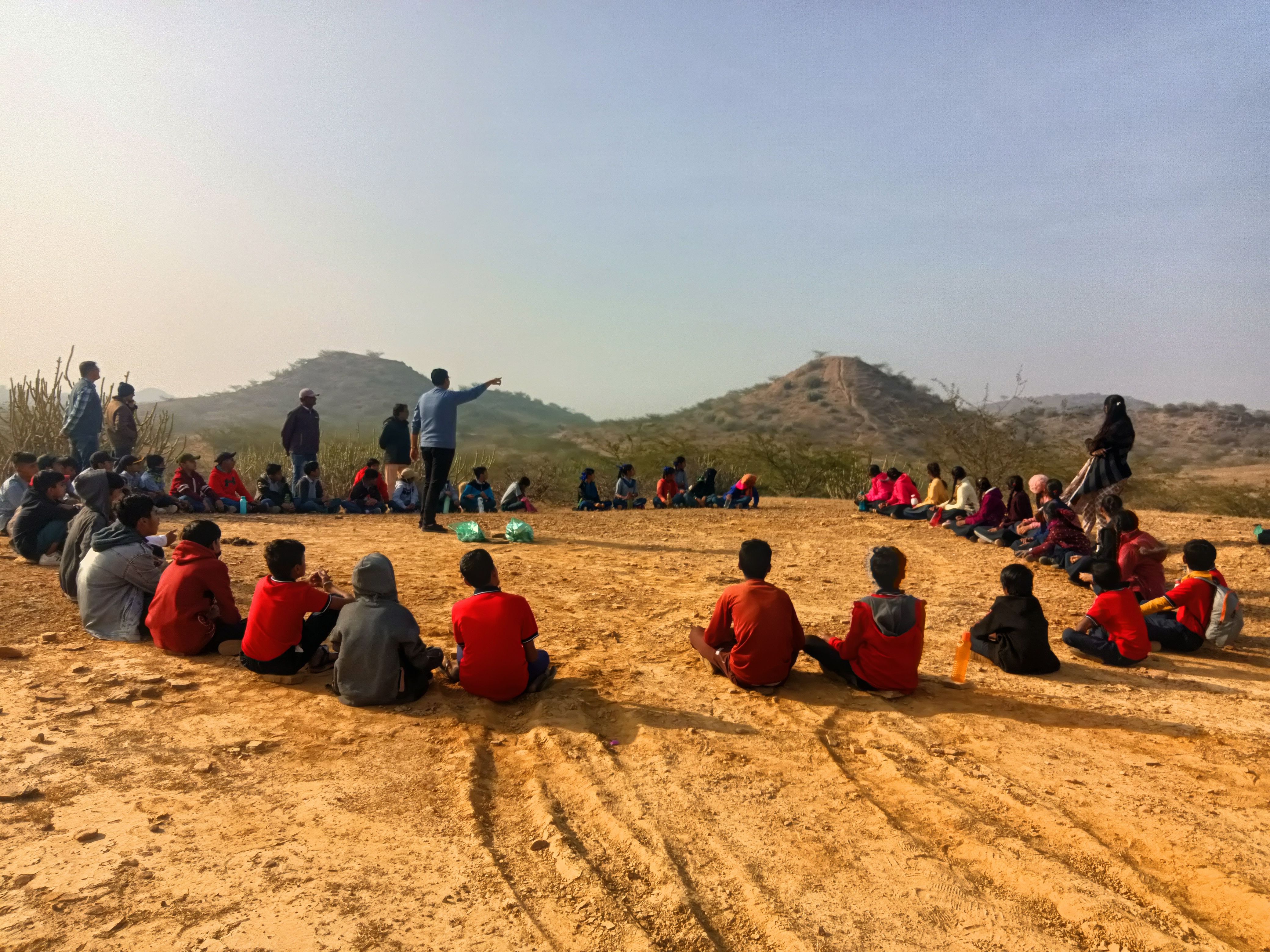 Children are sitting in a circle in an open space. A man is standing and pointing his hand towards a hill in the distance.