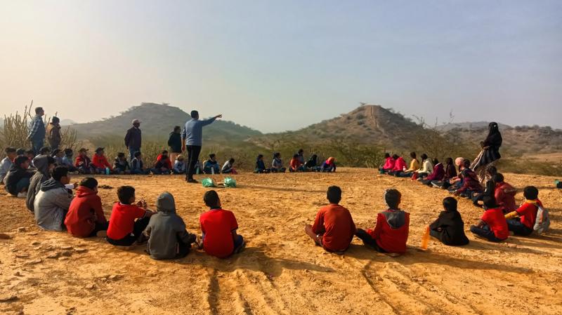 Children are sitting in a circle in an open space. A man is standing and pointing his hand towards a hill in the distance.